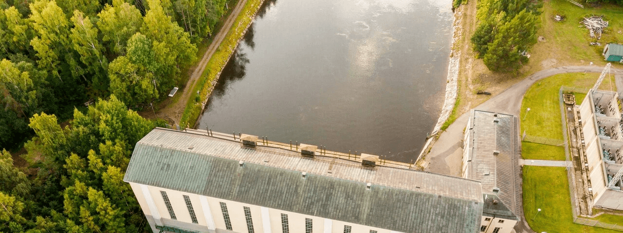 Drönarbild över ett vattenkraftverk där lugnt vatten flyter genom turbinhuset som är omgivet av grön skog.